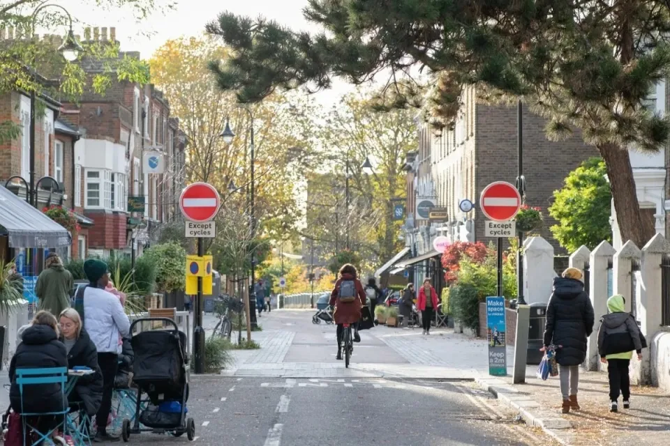 Person on bike on small road in London with people eating at outdoor dining tables and pedestrians walking on either side.